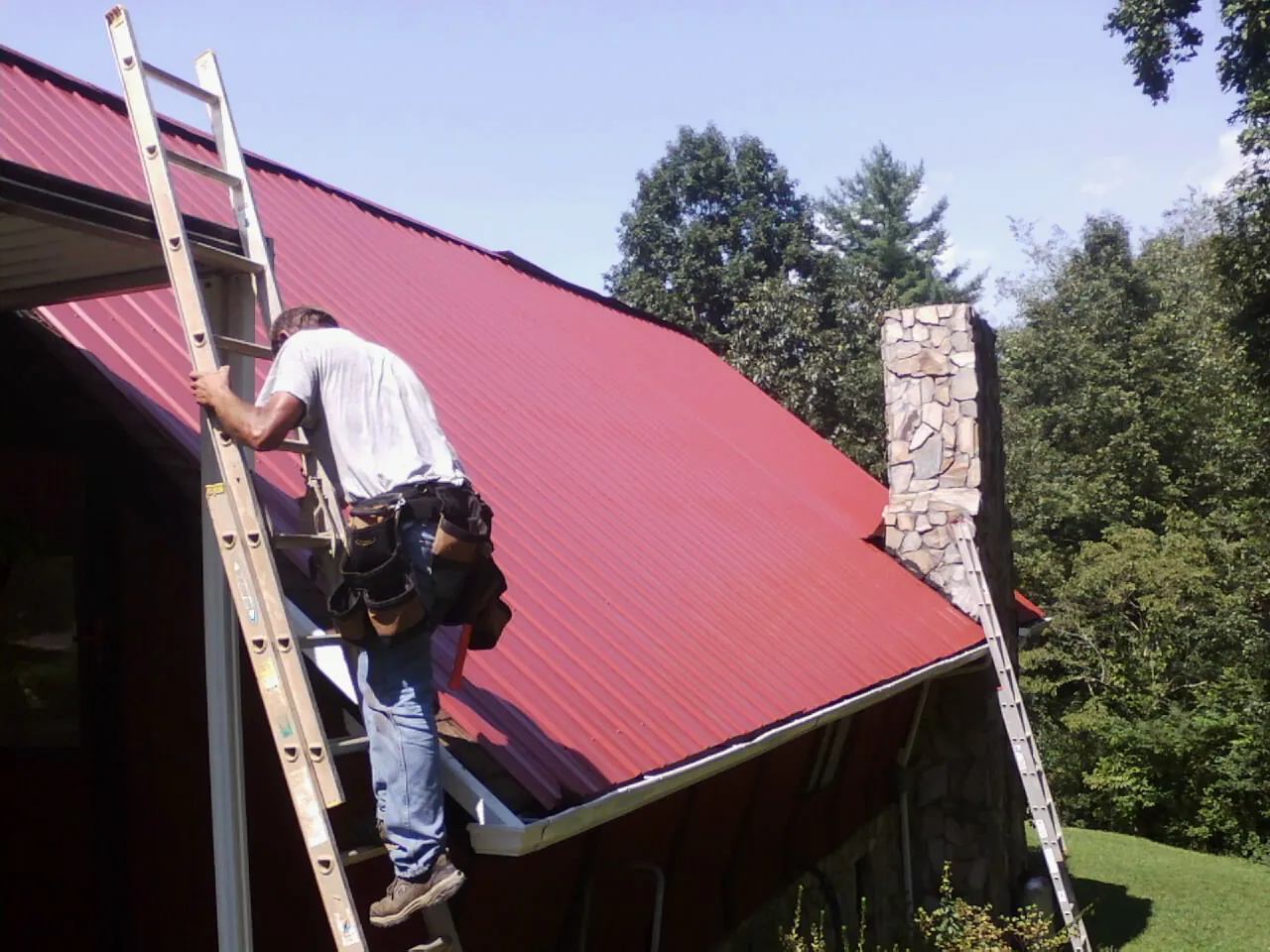 Roofer on a ladder beside a red metal roof and stone chimney — Soffit & Fascia Repair by Roofing Brothers in Poughkeepsie